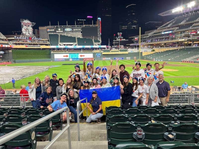 A group of international youth delegates and facilitators holding a Ukrainian flag at Target Field baseball stadium, smiling and united during a YouLEAD 2025 community event.
