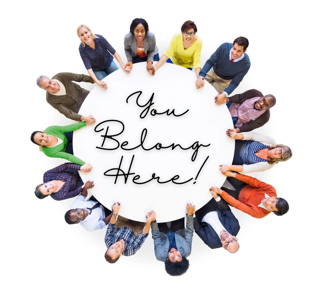 A diverse group of leaders holding hands in a circle around a table with the words “You Belong Here” in the center.